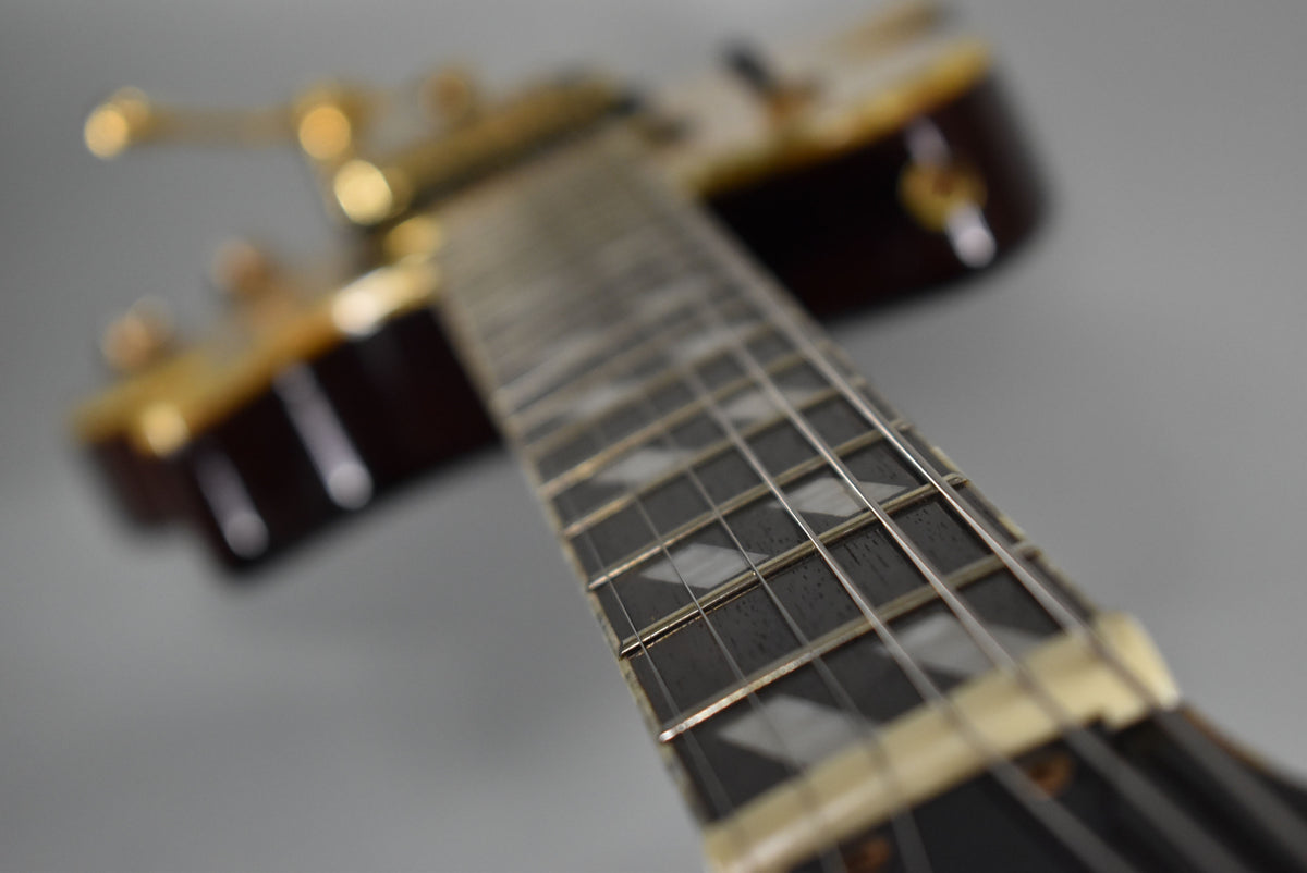 Close-up of the guitar's dark fretboard with diamond inlays, angled from the nut towards the blurred, gold-hardware headstock.