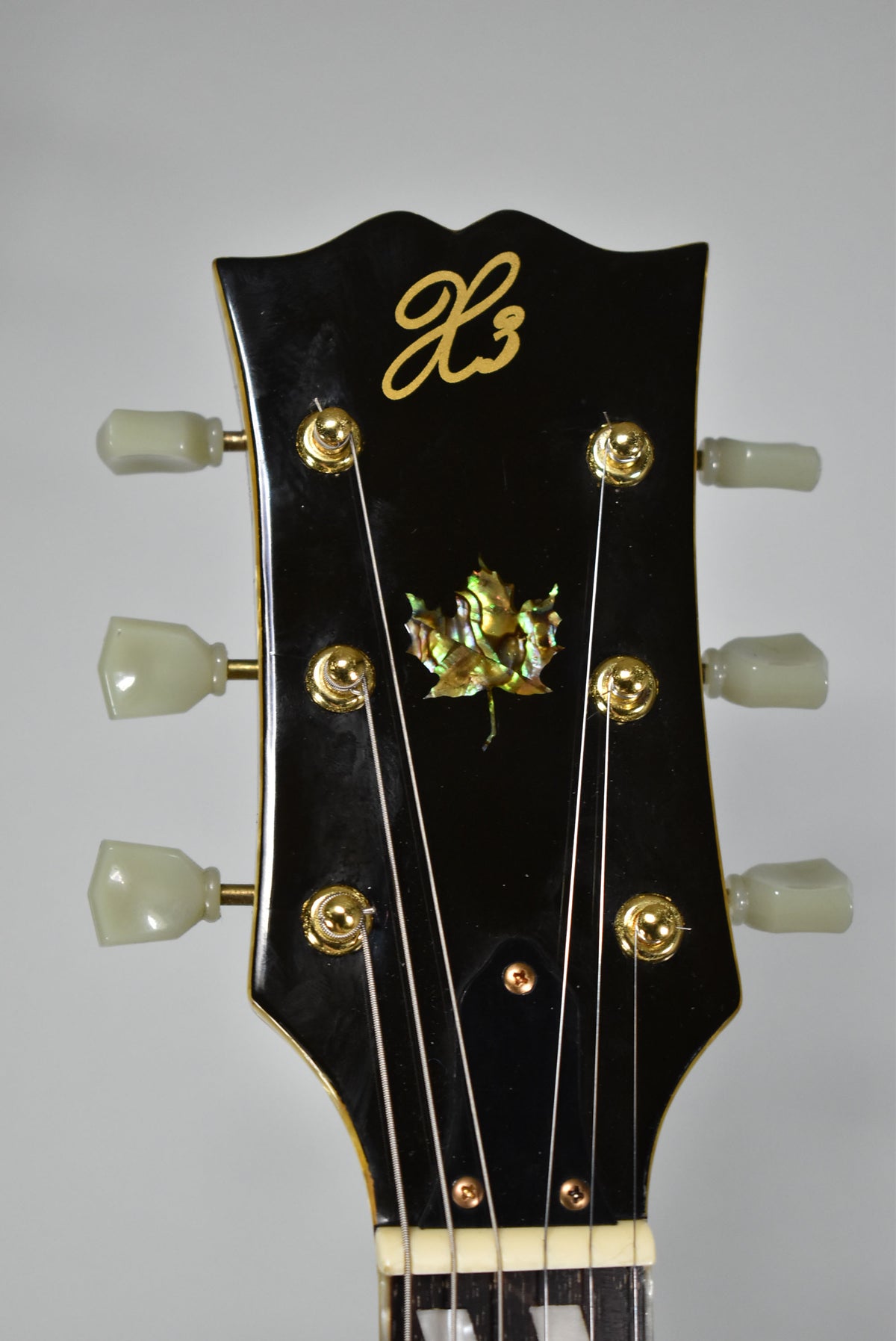 Close-up of the guitar's glossy black headstock with gold tuners, cream tuning pegs, and a mother-of-pearl maple leaf inlay.
