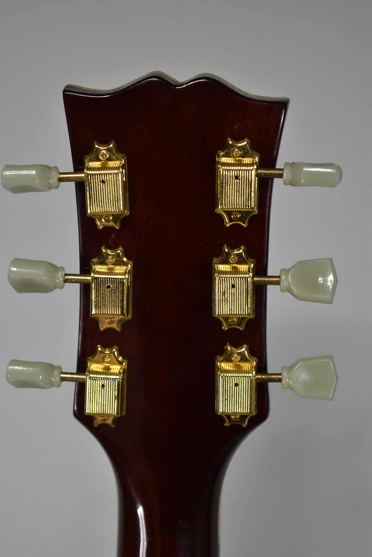 The back of the guitar's headstock, dark brown with a glossy finish, showing six gold-plated tuners with cream-colored buttons.