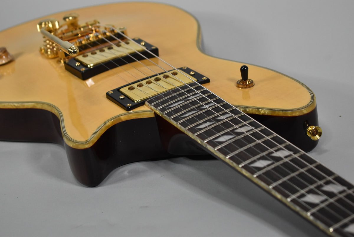 The guitar's natural maple top with gold hardware and a dark mahogany body is shown from the upper bout, angled towards the fretboard.